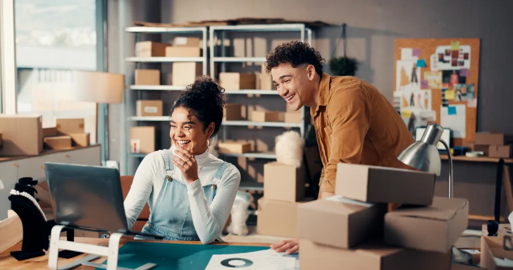 Two Gen Z small business owners share a laugh while reviewing a laptop together in their workspace surrounded by shipping boxes