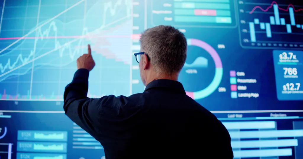 A man points at a large screen of graphs and data