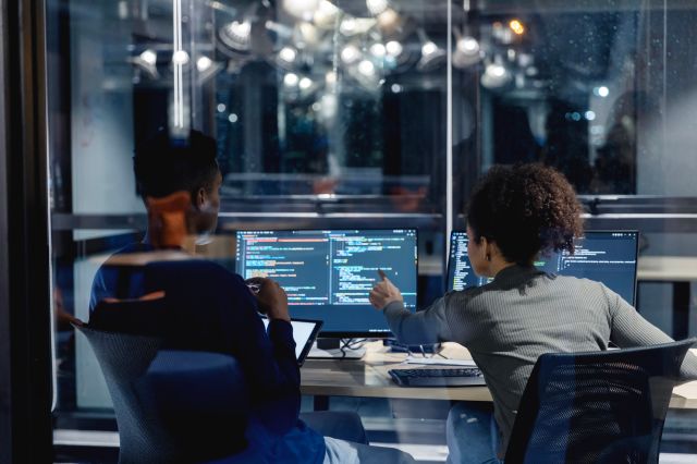 Two employees sit in chairs at a desk to review code on a computer screen.
