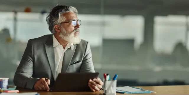Older male Chief Financial Officer holding an iPad at a desk and looking off in the distance.