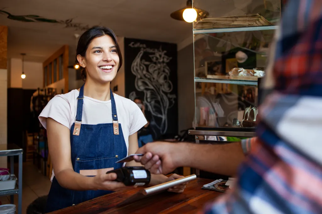 Smiling young female employee wearing a denim pinafore while holding a payment terminal at a coffee store in the morning