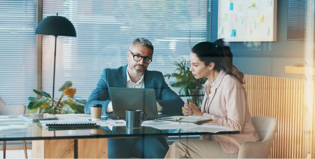 Man and woman sit at a desk in an office looking at corporate payments on a laptop.