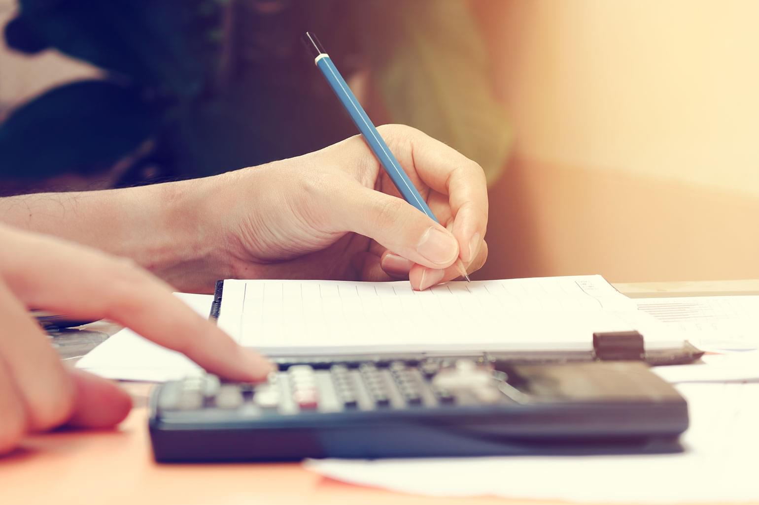 Plan rapproché d’une jeune femme qui fait des calculs chez elle, à l’aide d’une calculatrice, tout en prenant des notes à la main dans un cahier. Concept d’épargne et de finances.