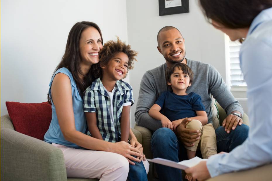 Une jeune famille de quatre personnes avec deux fils assis sur un canapé blanc, souriant pendant qu'un spécialiste en hypothèques discute du déménagement des hypothèques pour leur maison