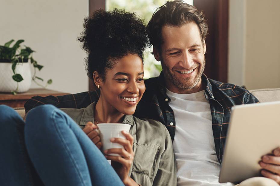 Image d'un couple heureux assis sur un sofa dans un moment de détente, qui regarde l'écran de la tablette que tient l'homme.