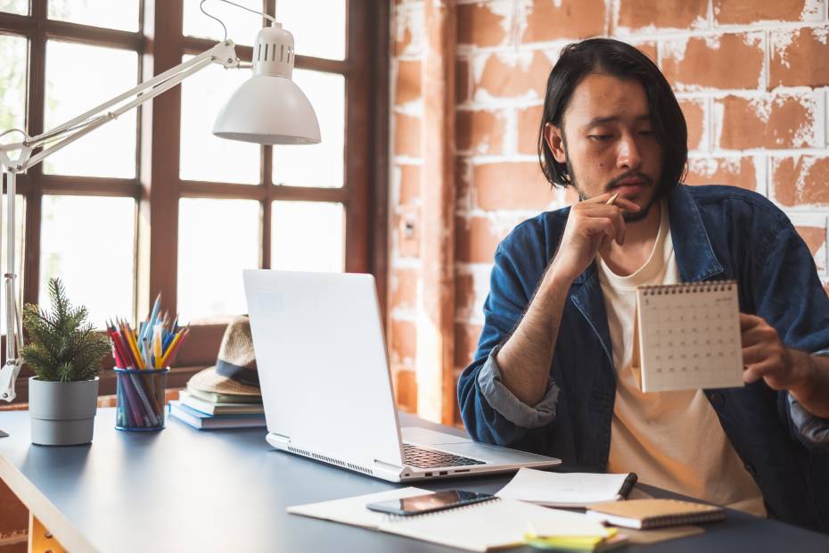 Jeune homme utilisant un ordinateur portable à un bureau et regardant un calendrier.