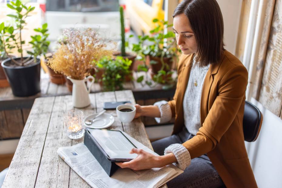 Une femme portant un blazer tient une chope à café et lit les informations sur une tablette sous laquelle un journal est déposé sur un bureau en bois.