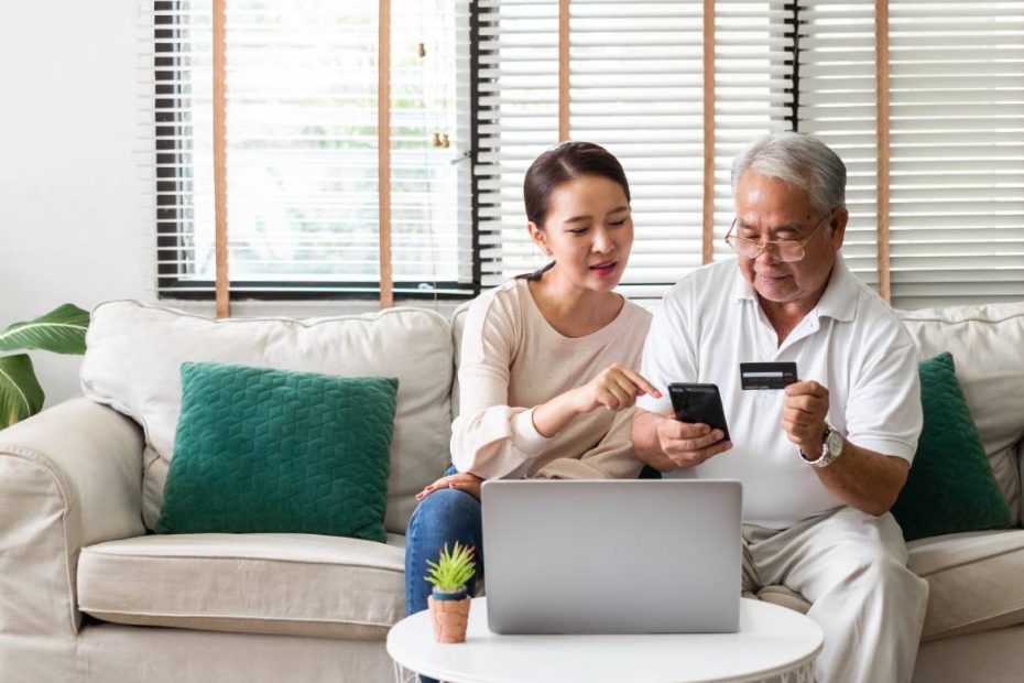 Une femme asiatique explique à un homme plus âgé comment utiliser une carte de crédit et un téléphone pour faire des achats en ligne.