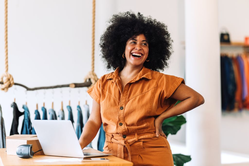 Propriétaire d'une petite entreprise ethnique souriant joyeusement tout en se tenant dans son magasin. Heureuse femme d'affaires gérant ses commandes de vêtements sur un ordinateur portable. Femme entrepreneur noire qui gère une boutique de vêtements en ligne.