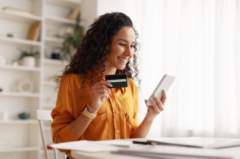 Femme arabe joyeuse utilisant un smartphone faisant des achats en ligne tenant une carte de crédit effectuant un paiement assise au bureau à la maison.