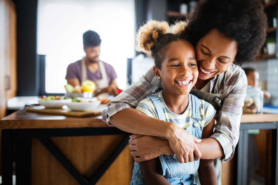 Une mère souriante enlace sa fille qui rit dans une cuisine chaleureuse, tandis qu’une autre personne prépare de la nourriture à l’arrière-plan.