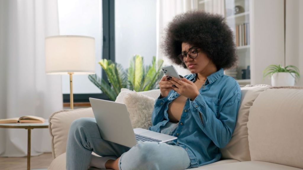 Jeune femme assise sur un canapé, un ordinateur sur les genoux et un téléphone à la main.
