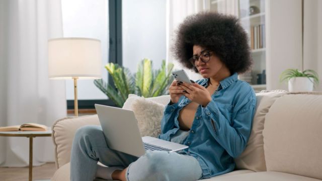 Young woman sitting on a couch with a computer on her lap and a phone in her hands.