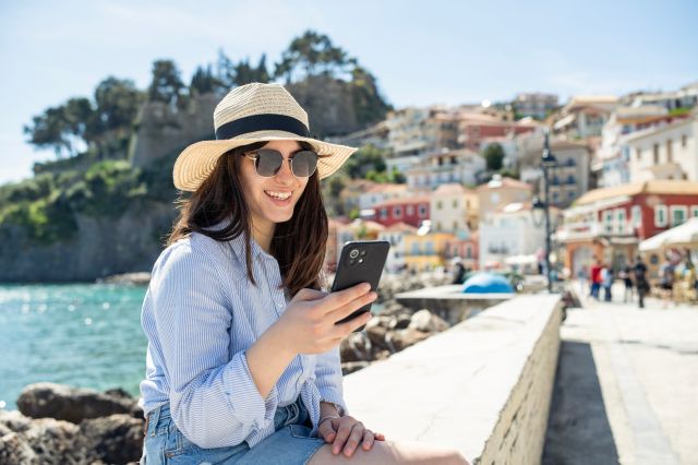 Une femme souriante portant un chapeau de soleil et des lunettes de soleil utilise son téléphone intelligent assise sur une promenade côtière ensoleillée, avec une ville balnéaire méditerranéenne colorée en arrière-plan.