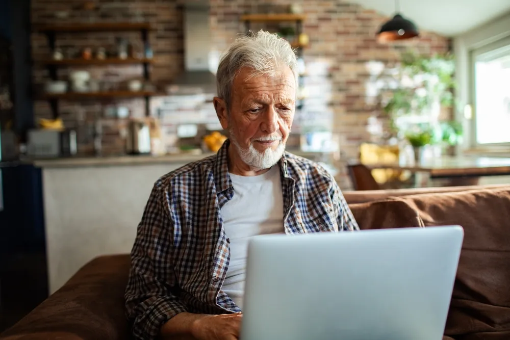 Elderly man sitting on a couch while browsing on his computer.