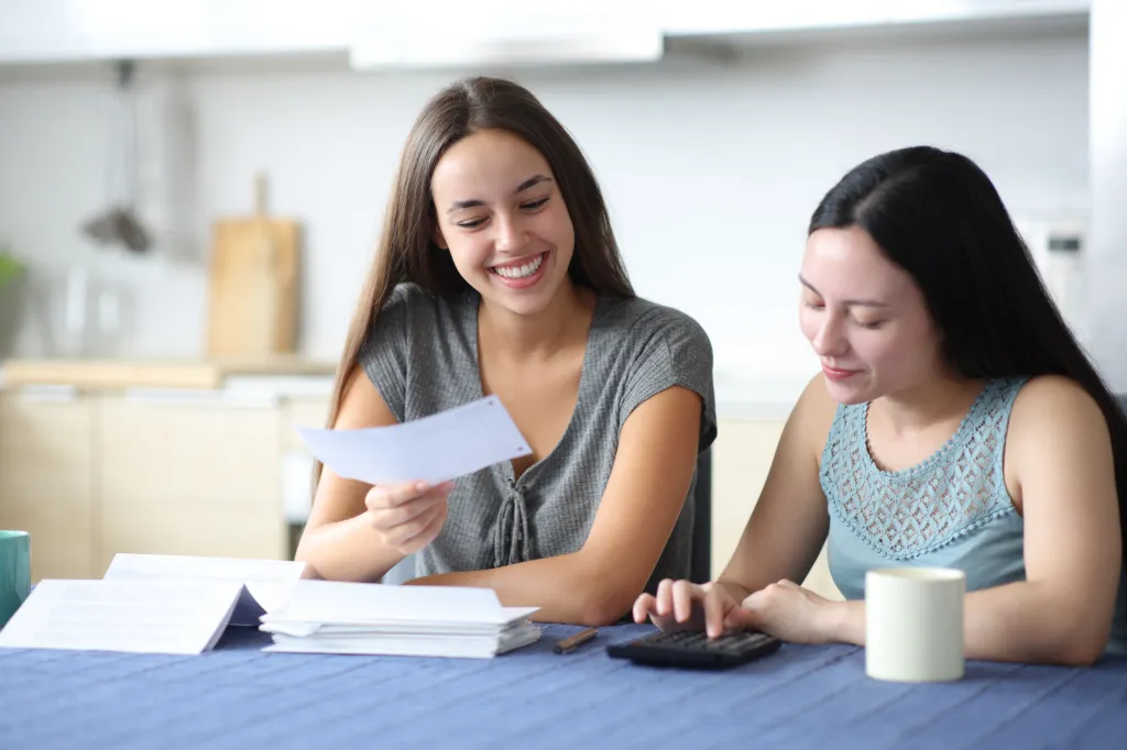 Deux personnes à une table de cuisine nappée de bleu examinent des documents ; l'une sourit en tenant un papier, l'autre utilise une calculatrice.