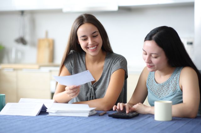 Deux personnes à une table de cuisine nappée de bleu examinent des documents ; l'une sourit en tenant un papier, l'autre utilise une calculatrice.